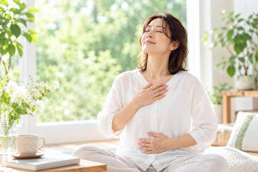 窓辺で目を閉じて手を心臓とお腹に当て、呼吸を感じている穏やかな雰囲気の女性